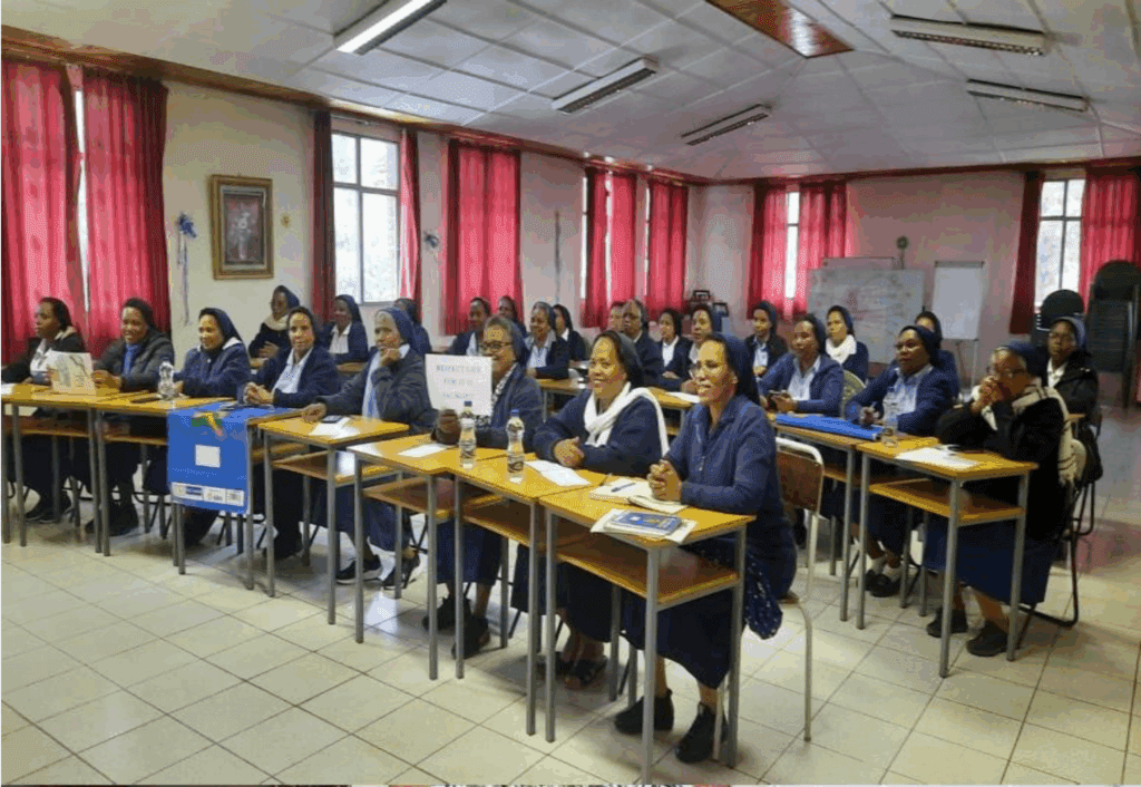 In einem Klassenraum mit roten Vorhängen sitzen etwa zwanzig Ordensschwestern in blauen Gewändern und weißen Schleiern aufmerksam an Schultischen. Einige halten Schilder hoch, darunter eines mit der Aufschrift „REFLECTIVE PRACTICE FOR RELIGIOUS TEACHERS“. Auf den Tischen liegen Wasserflaschen, Notizhefte und Informationsmaterial. Die Stimmung wirkt konzentriert und freundlich.