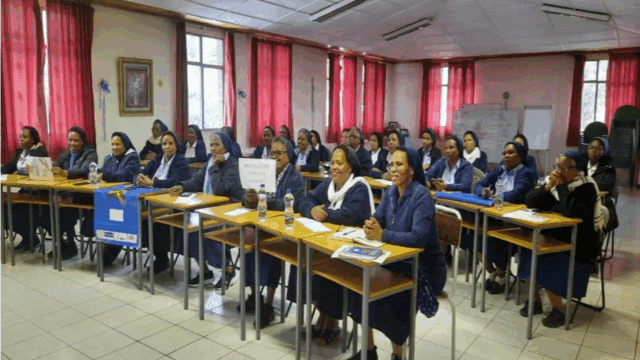 In einem Klassenraum mit roten Vorhängen sitzen etwa zwanzig Ordensschwestern in blauen Gewändern und weißen Schleiern aufmerksam an Schultischen. Einige halten Schilder hoch, darunter eines mit der Aufschrift „REFLECTIVE PRACTICE FOR RELIGIOUS TEACHERS“. Auf den Tischen liegen Wasserflaschen, Notizhefte und Informationsmaterial. Die Stimmung wirkt konzentriert und freundlich.