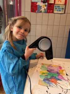 Ein junges Mädchen in einem blauen Kittel lächelt, während es an einem Tisch in einem Klassenzimmer ein buntes Aquarell mit einem schwarzen Föhn trocknet.