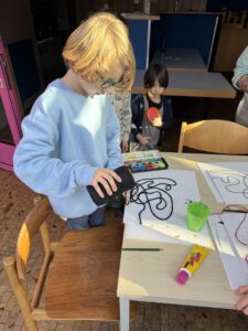 Ein Kind mit blondem Haar und Brille schüttet schwarze Schnur auf Papier und schafft so ein abstraktes Muster, während ein anderes Kind im Hintergrund einen roten Deckel hält. Kunstzubehör und bunte Farben liegen auf dem Tisch im Sonnenlicht.