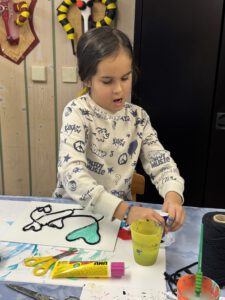 Ein kleines Kind sitzt an einem Tisch und arbeitet an einem Kunstprojekt mit einer schwarz umrandeten Zeichnung eines Pandas in Weiß und Grüntönen. Kleber, Schere und Farbe liegen verstreut herum.