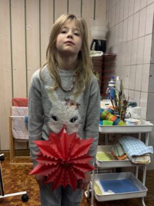 Ein kleines Kind mit langen blonden Haaren, das einen grauen Pullover mit einem Tiergesicht trägt, steht in einem Haus und hält einen roten Papierstern in der Hand. In den Regalen hinter ihnen sind Malutensilien und Handtücher zu sehen.