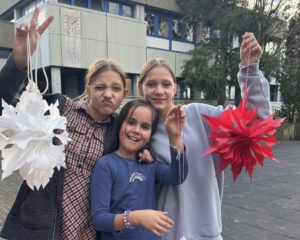 Drei lächelnde Kinder stehen draußen und halten selbstgebastelte weiße und rote Papierlaternen. Ein Kind macht ein verspieltes Gesicht und ein Friedenszeichen. Im Hintergrund sind Bäume und ein Gebäude mit blau umrandeten Fenstern zu sehen.