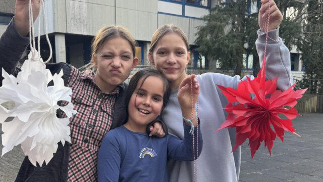Drei lächelnde Kinder stehen draußen und halten selbstgebastelte weiße und rote Papierlaternen. Ein Kind macht ein verspieltes Gesicht und ein Friedenszeichen. Im Hintergrund sind Bäume und ein Gebäude mit blau umrandeten Fenstern zu sehen.